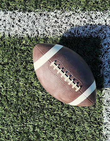 A close-up of a football placed on a grassy end zone line, casting a shadow under bright sunlight.