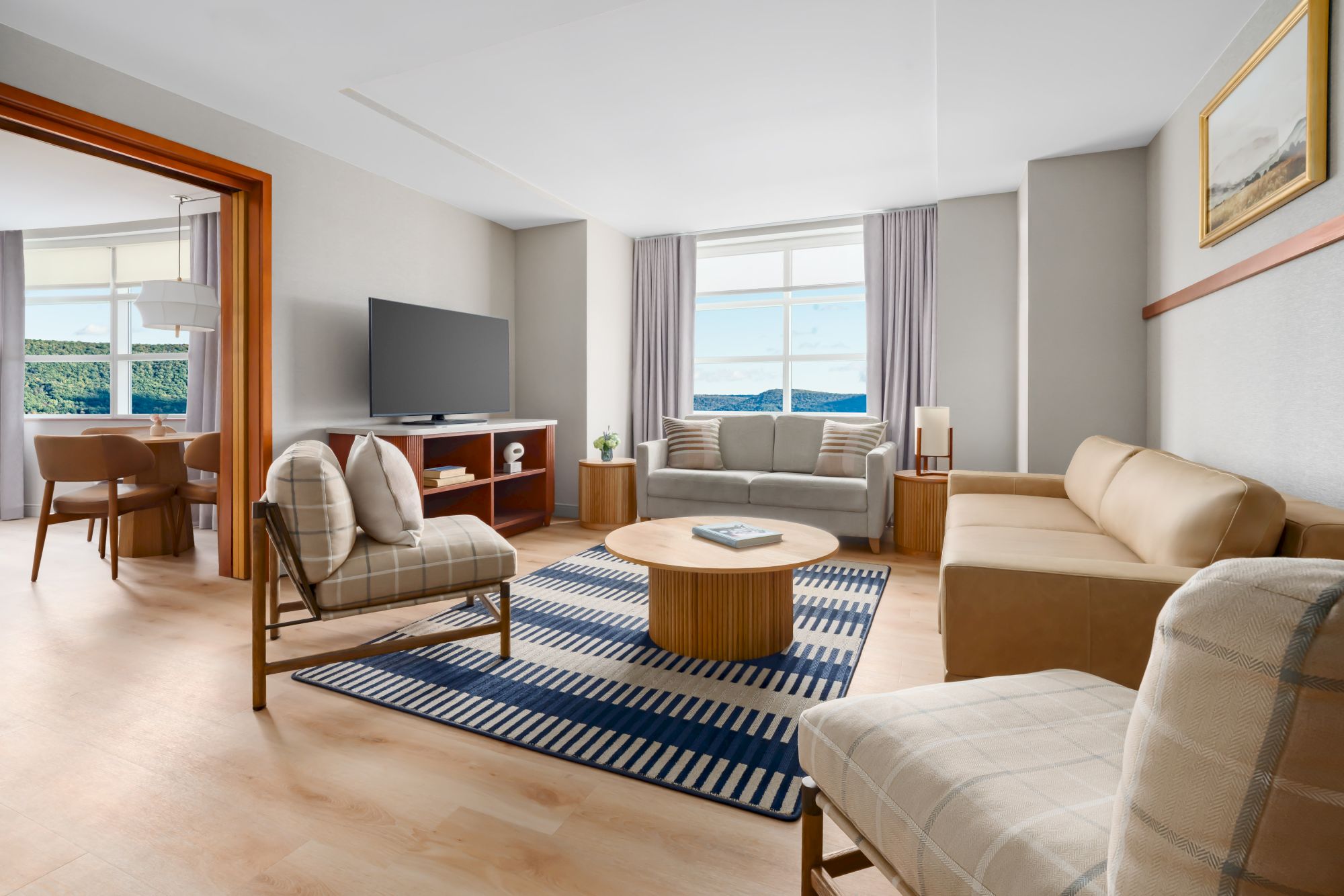 Bright modern living room with beige sofas, a TV on a stand, coffee table, rugs, and large windows.