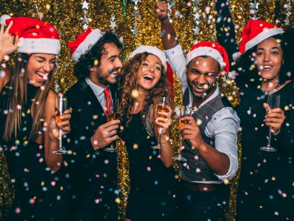 A group of friends in Santa hats cheer and toast champagne at a festive party as confetti sparkles.
