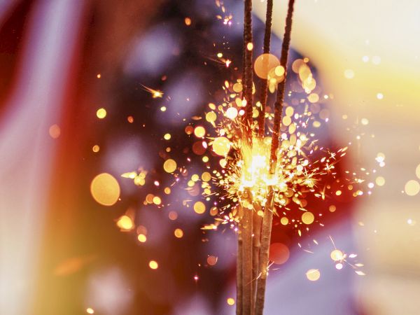 A close-up of glowing incense sticks with bright sparks and drifting embers amid warm bokeh lights.