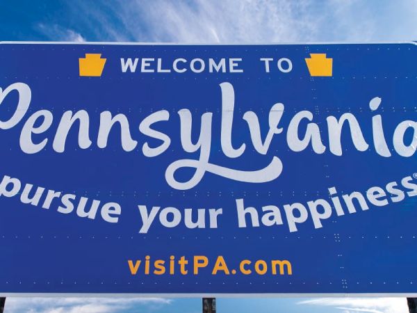 A blue &ldquo;Welcome to Pennsylvania&rdquo; roadside sign says &ldquo;Pursue your happiness&rdquo; with &ldquo;visitPA.com&rdquo; under it.