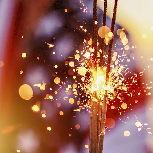 A glowing sparkler or firework shoots bright sparks through the air, with colorful bokeh lights in the background.