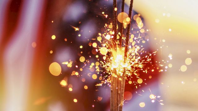 A glowing sparkler or firework shoots bright sparks through the air, with colorful bokeh lights in the background.