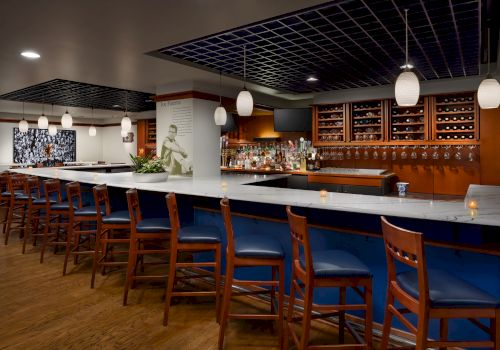 A modern bar interior with a marble counter, bottle shelves, hanging lantern lights, and wooden stools.