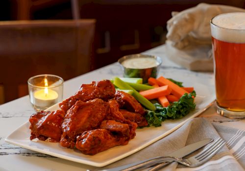 A plated meal of saucy meat with fresh carrots, celery, broccoli, and greens, set with candles and a glass of beer on a table.