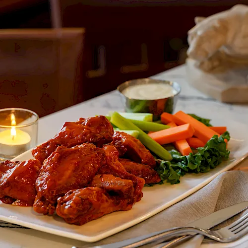 A plated meal of saucy meat with fresh carrots, celery, broccoli, and greens, set with candles and a glass of beer on a table.