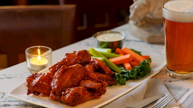 A plated meal of saucy meat with fresh carrots, celery, broccoli, and greens, set with candles and a glass of beer on a table.