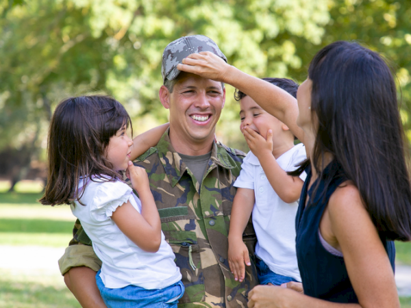 A smiling man in camouflage plays with three kids in a sunny park, children touching his hat and face.