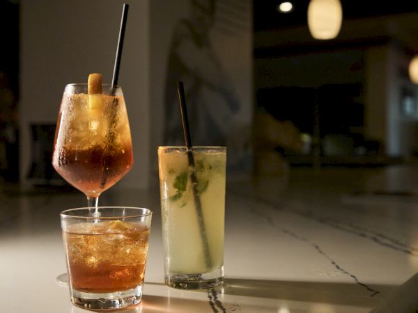 A bartender pours a drink into a glass with ice and a straw, while another tall cocktail sits beside it.
