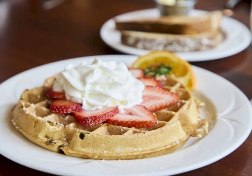 A plate of Belgian waffles topped with whipped cream and sliced strawberries, with a side of orange slices in the background.
