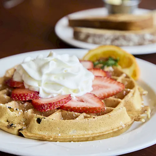 A plate of Belgian waffles topped with whipped cream and sliced strawberries, with a side of orange slices in the background.