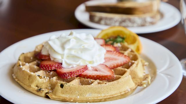 A plate of Belgian waffles topped with whipped cream and sliced strawberries, with a side of orange slices in the background.