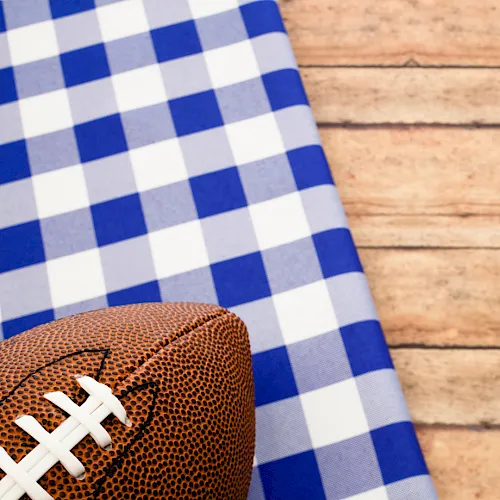 A brown football with white laces rests on blue-and-white checkered fabric against a wooden background.