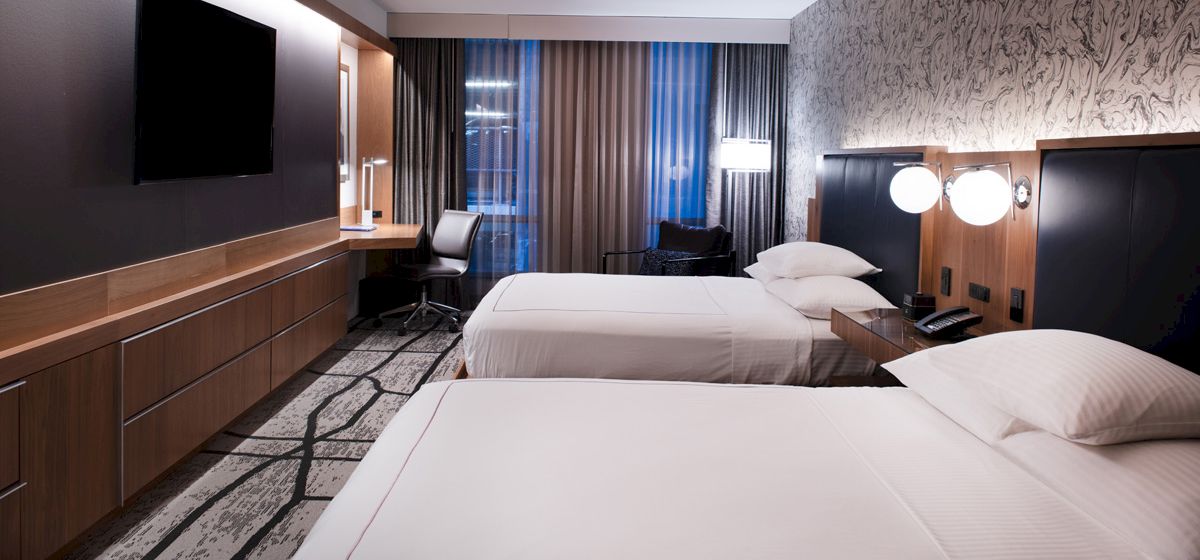 A modern hotel room with two neatly made beds, a desk chair, and a TV, lit by warm wall lamps.