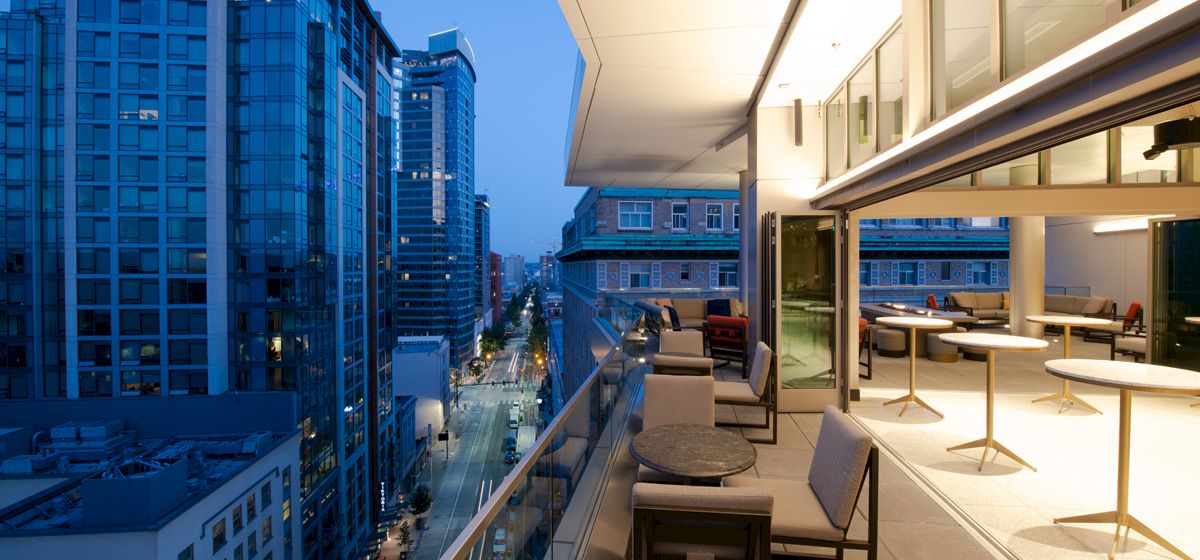 A high-rise balcony overlooks a downtown street with modern glass buildings and outdoor lounge seating at dusk.
