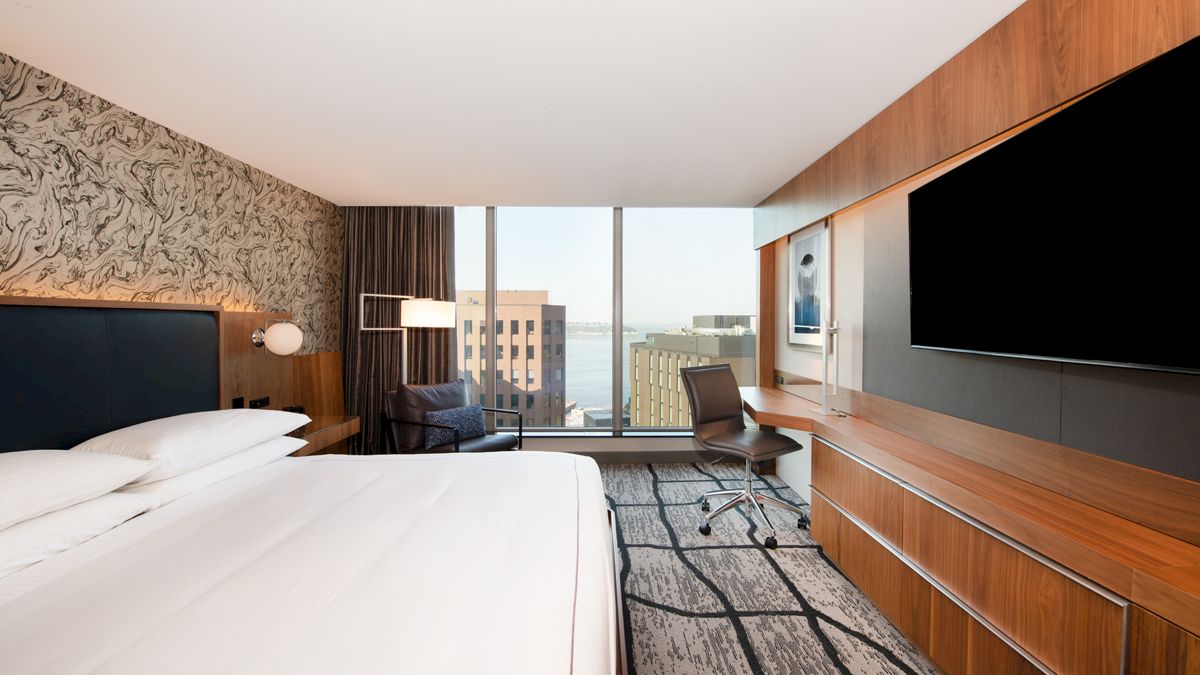 A modern hotel room featuring a large bed, desk chair, wall-mounted TVs, and floor-to-ceiling windows overlooking buildings.