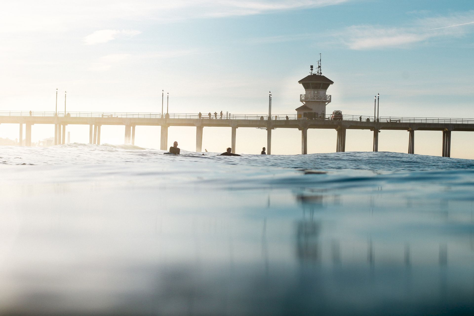 Sunlit pier over choppy water, with swimmers near the supports and a small control/watchtower on the deck.